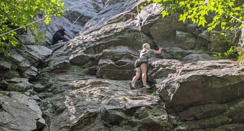 Two people wearing safety gear are secured by ropes as they each climb a rock face. 